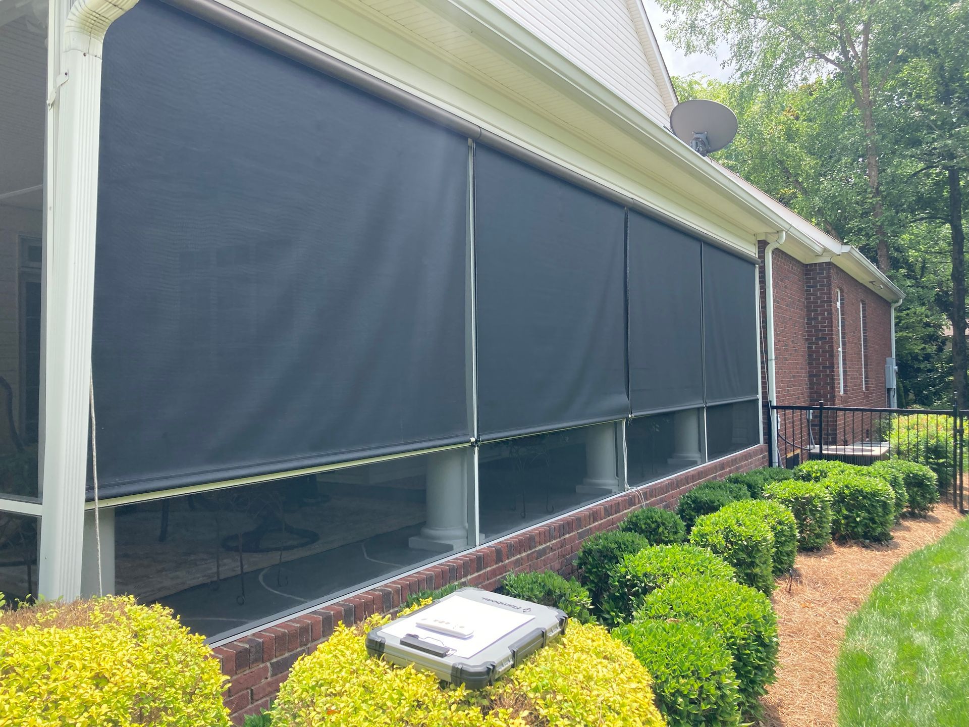 Black retractable sun shades on a brick house porch. Green bushes and a lawn in front.