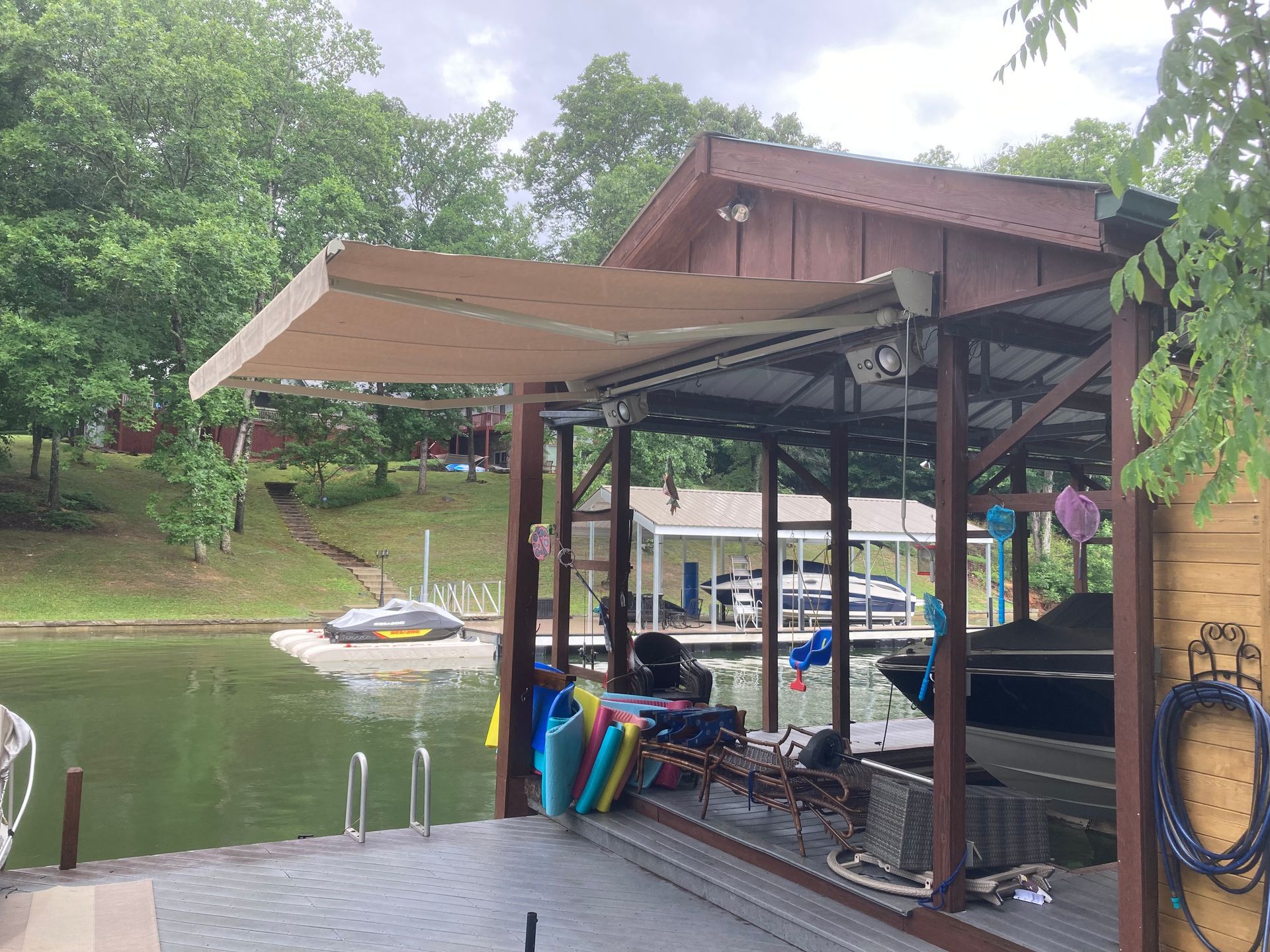Dock with boat lift, boat, and retractable beige awning over water. Trees and shoreline in background.
