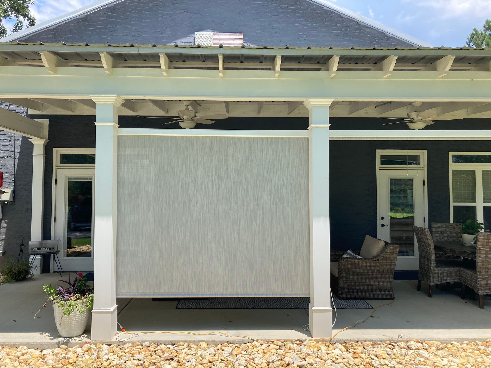 Outdoor patio with a retractable gray screen, supported by white columns, in front of a dark-colored house.