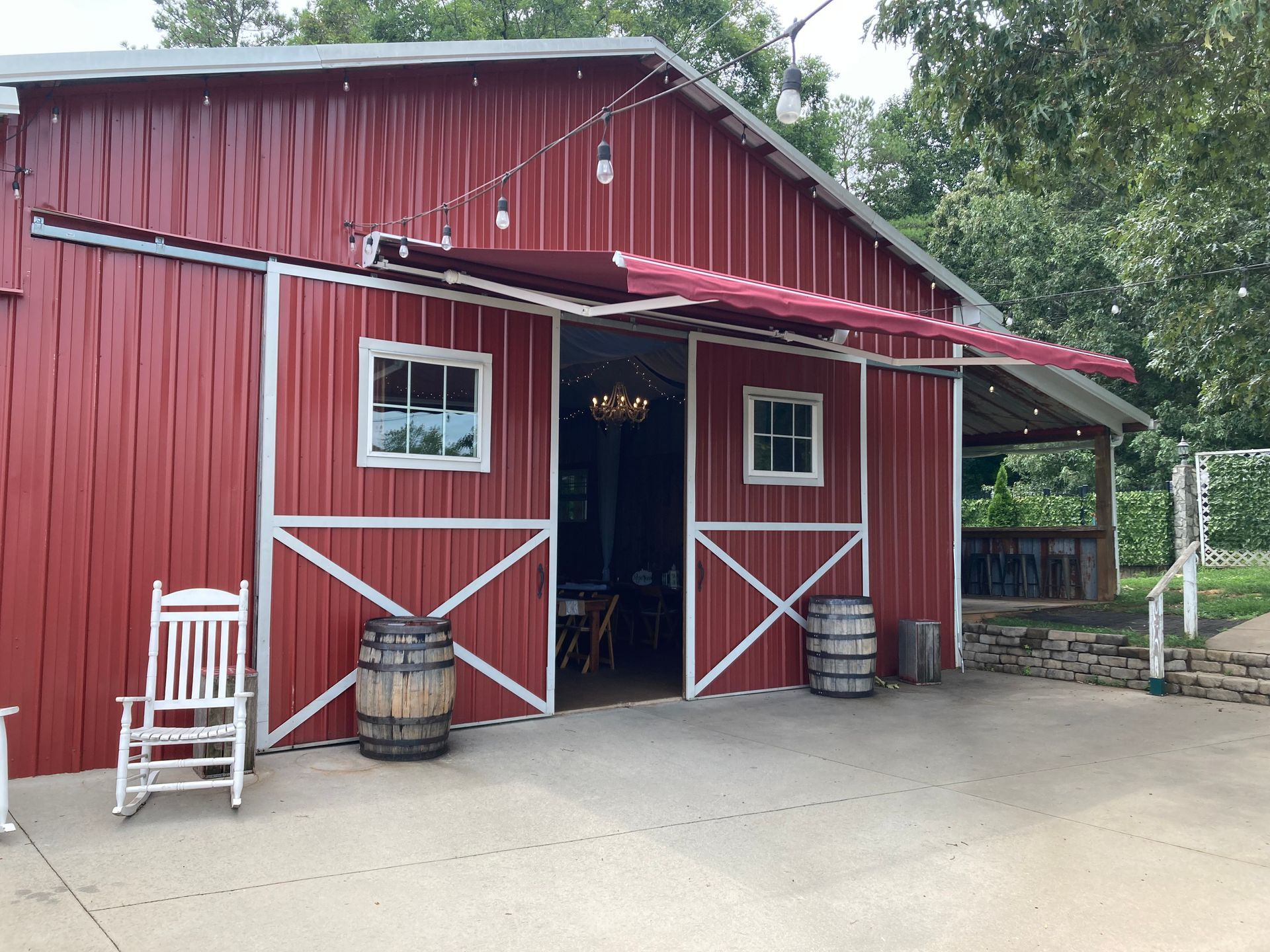 Red barn with white trim, open doors, and awning. Wooden barrels and a rocking chair sit outside.