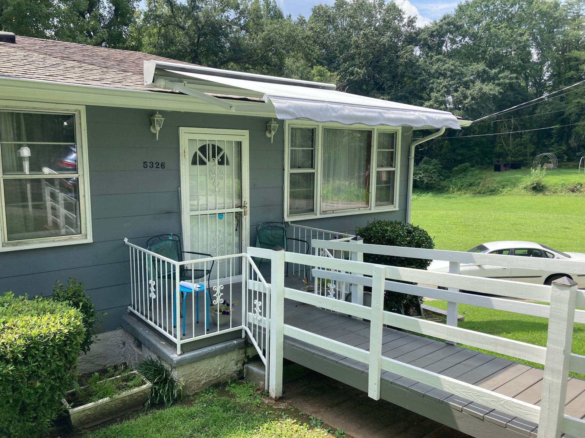 Gray house with a white ramp and railing. Green lawn, trees, and a car in the background.