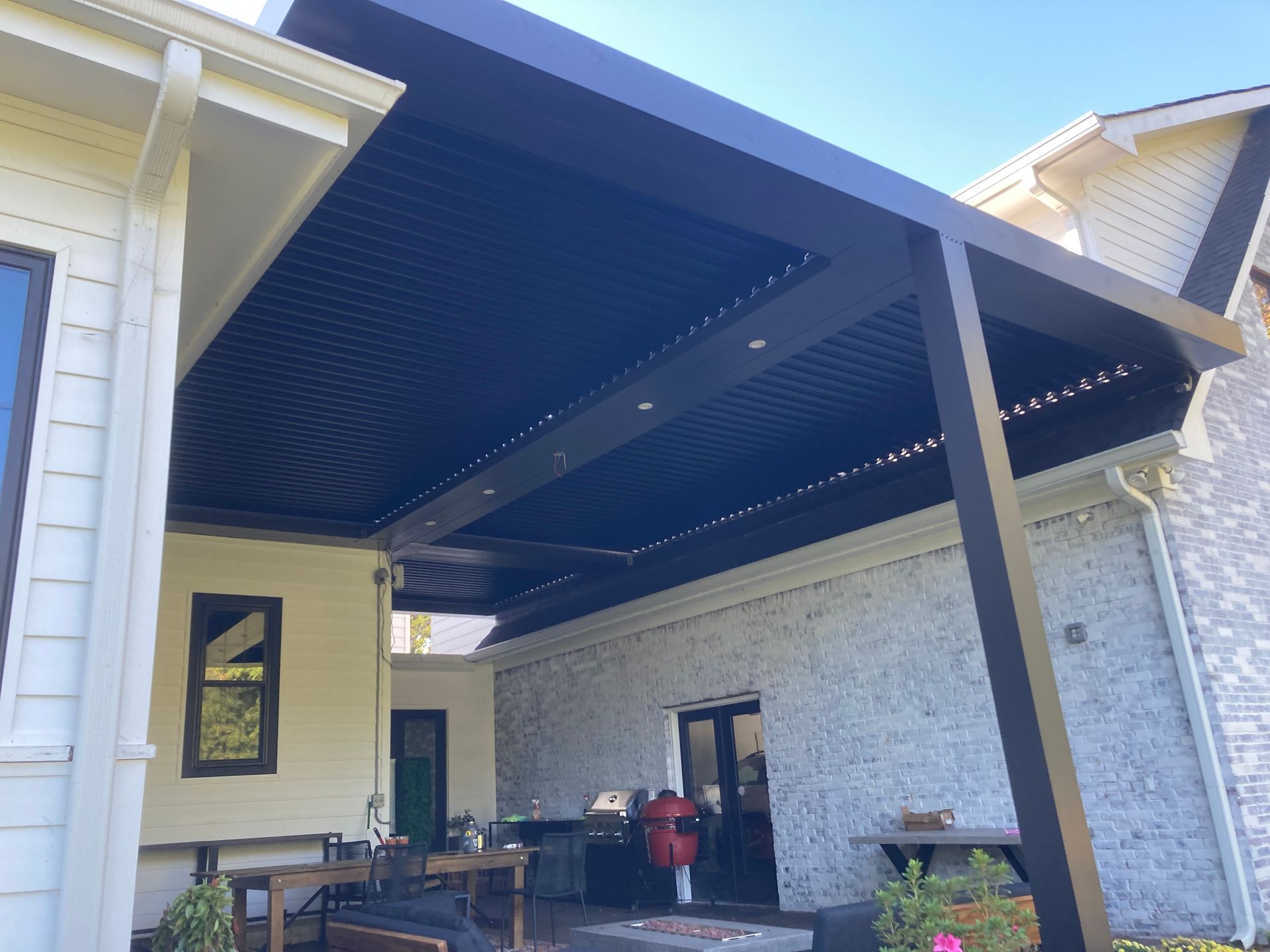Black pergola attached to a light-colored house, covering a patio area with built-in lights.