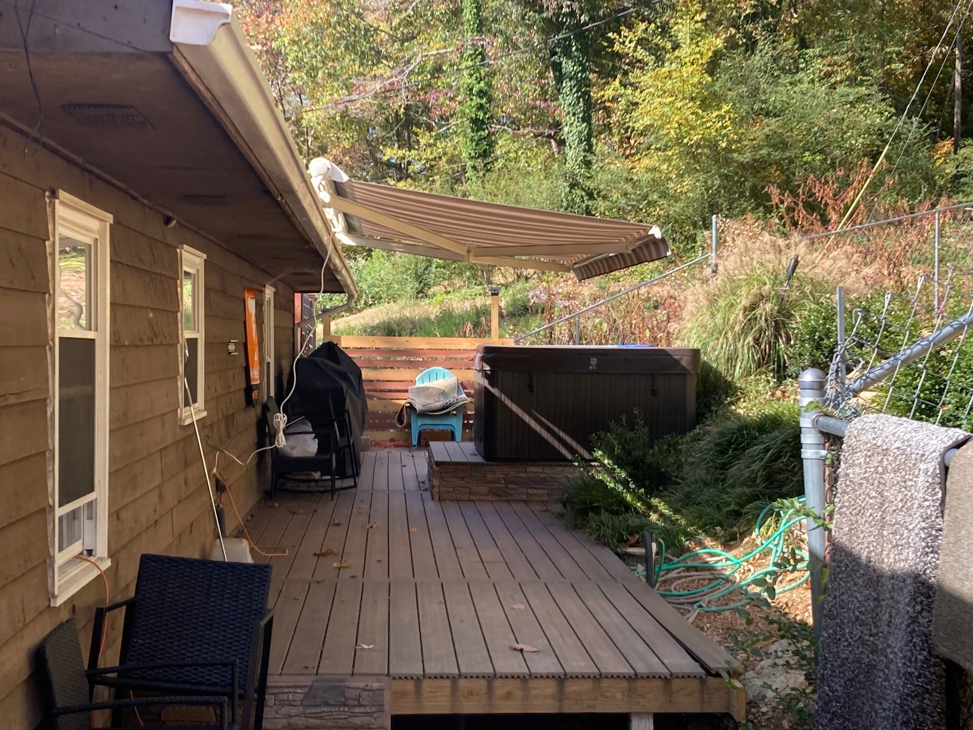 Wooden deck with a hot tub and an awning, beside a house with a wooded backdrop.
