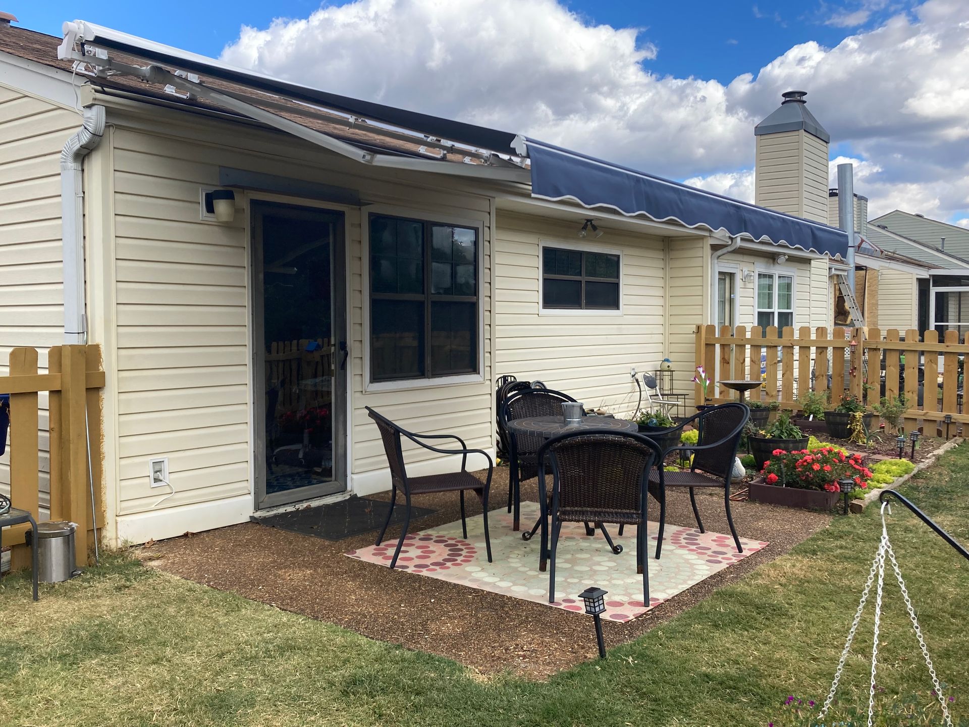 Backyard patio with table, chairs, plants, and a partial view of a house with beige siding, fence, and blue awning.