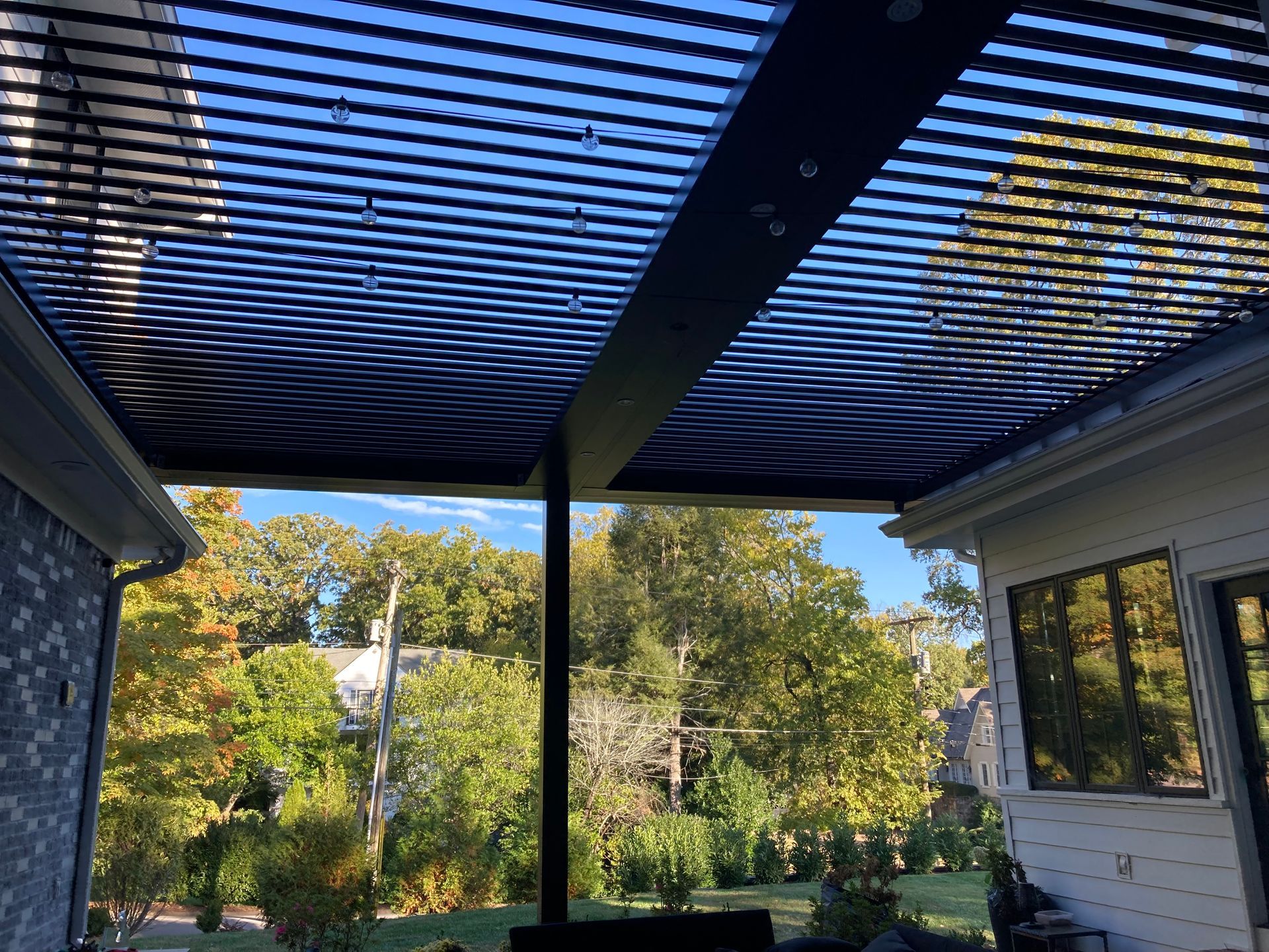 Outdoor patio roof with adjustable louvers, overlooking a yard with trees.