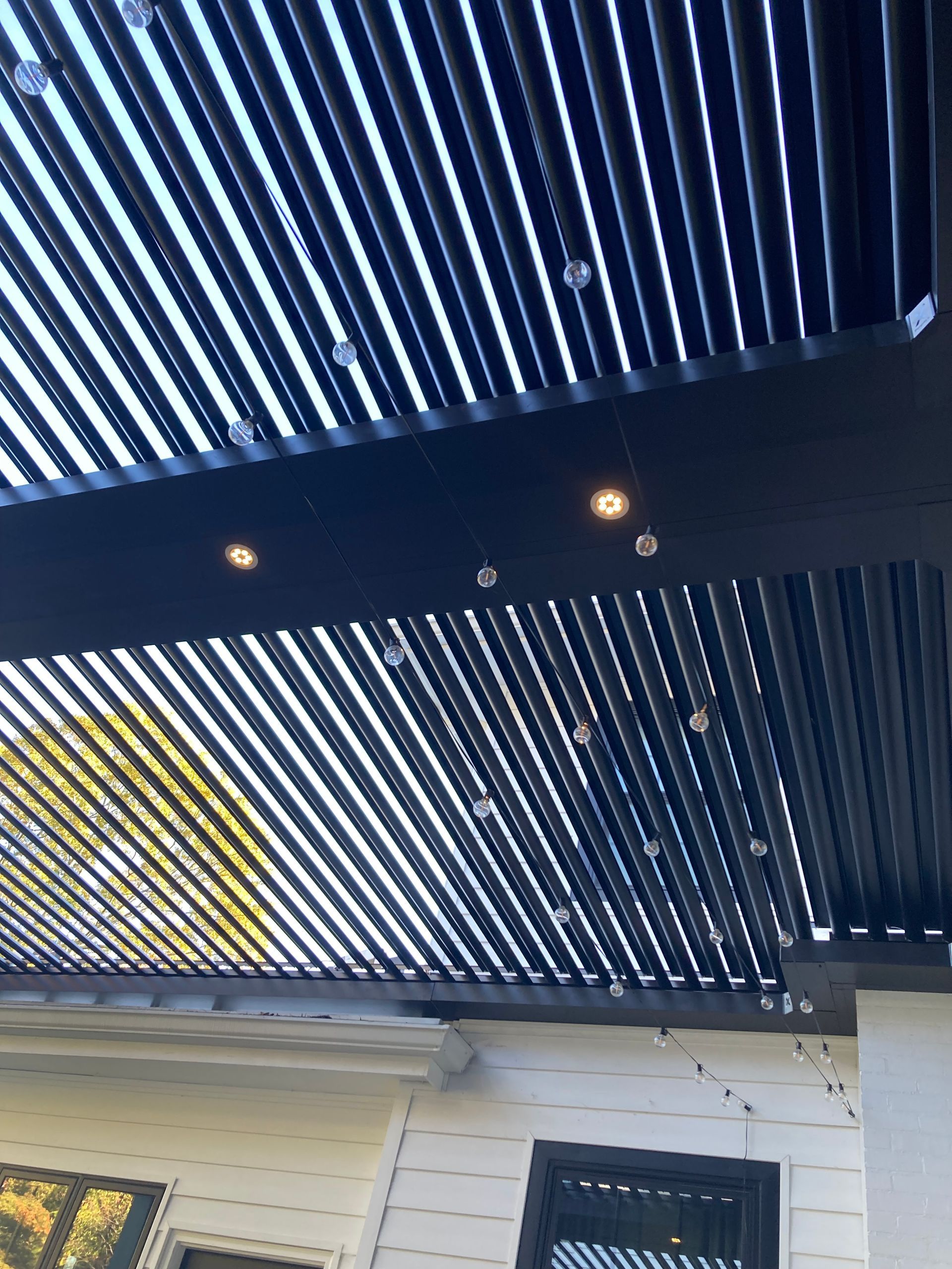 Black pergola roof with string lights, viewed from below against a bright sky.
