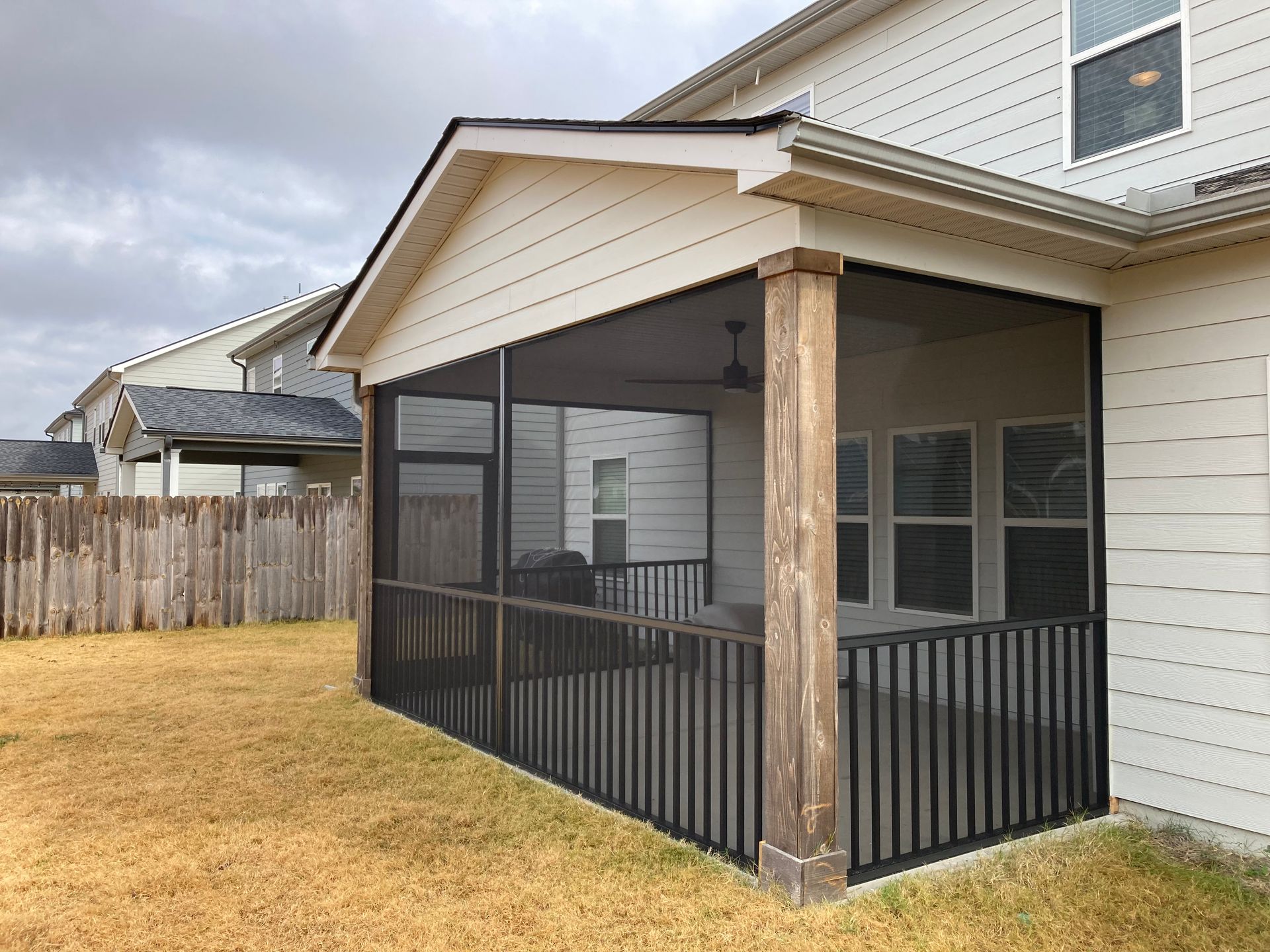 Screened-in porch on a house with brown wooden posts, black railings, and screen walls, overlooking a yard with brown grass.