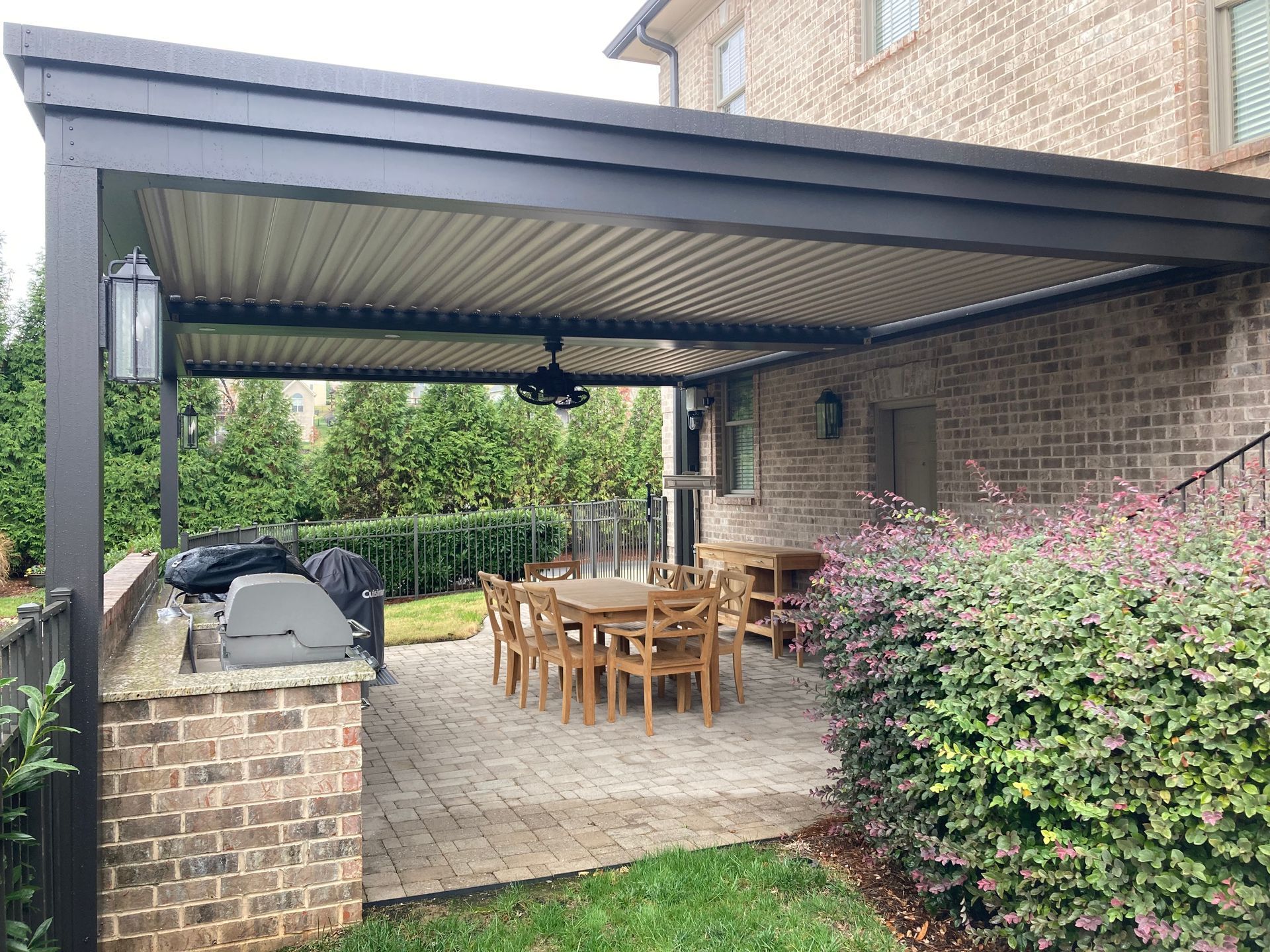 Patio with brick and stone, overhead covered seating, table, grill, and bushes.