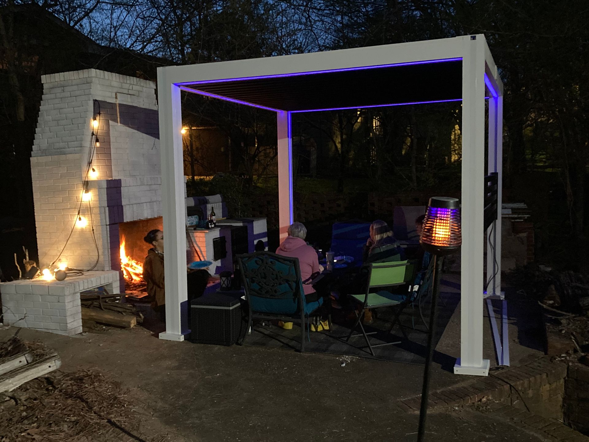 People seated under a white pergola with purple lights, near a lit fireplace.