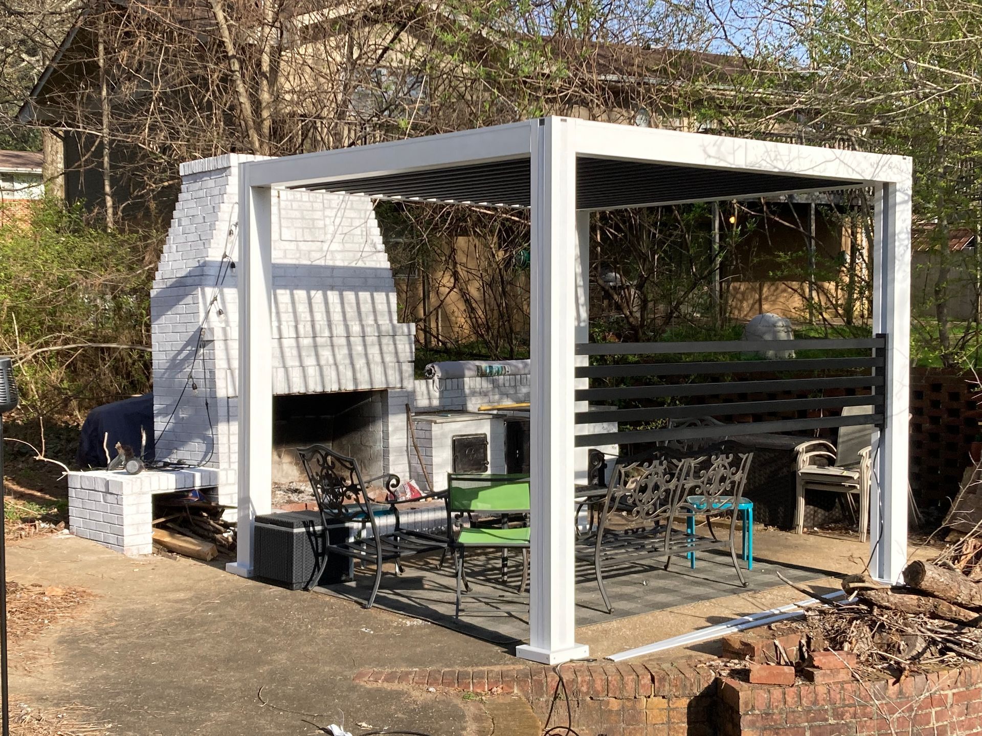 White pergola with grill and seating in a yard, trees in the background.