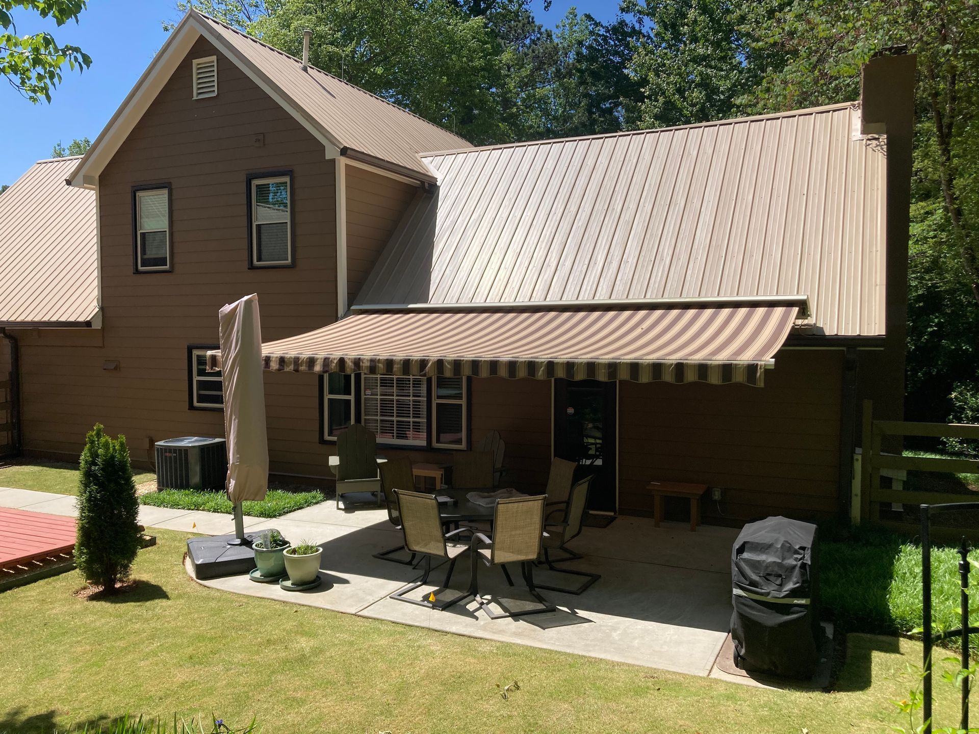 Brown house with metal roof, patio with awning, table, chairs, grill, and lawn.