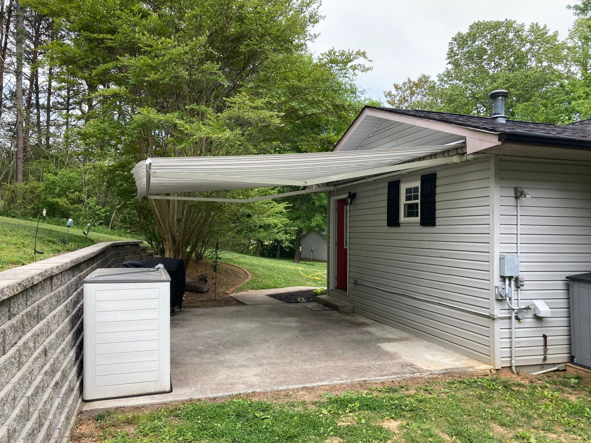 Patio with extending awning on a gray siding building, storage bin, brick wall, and green grass.