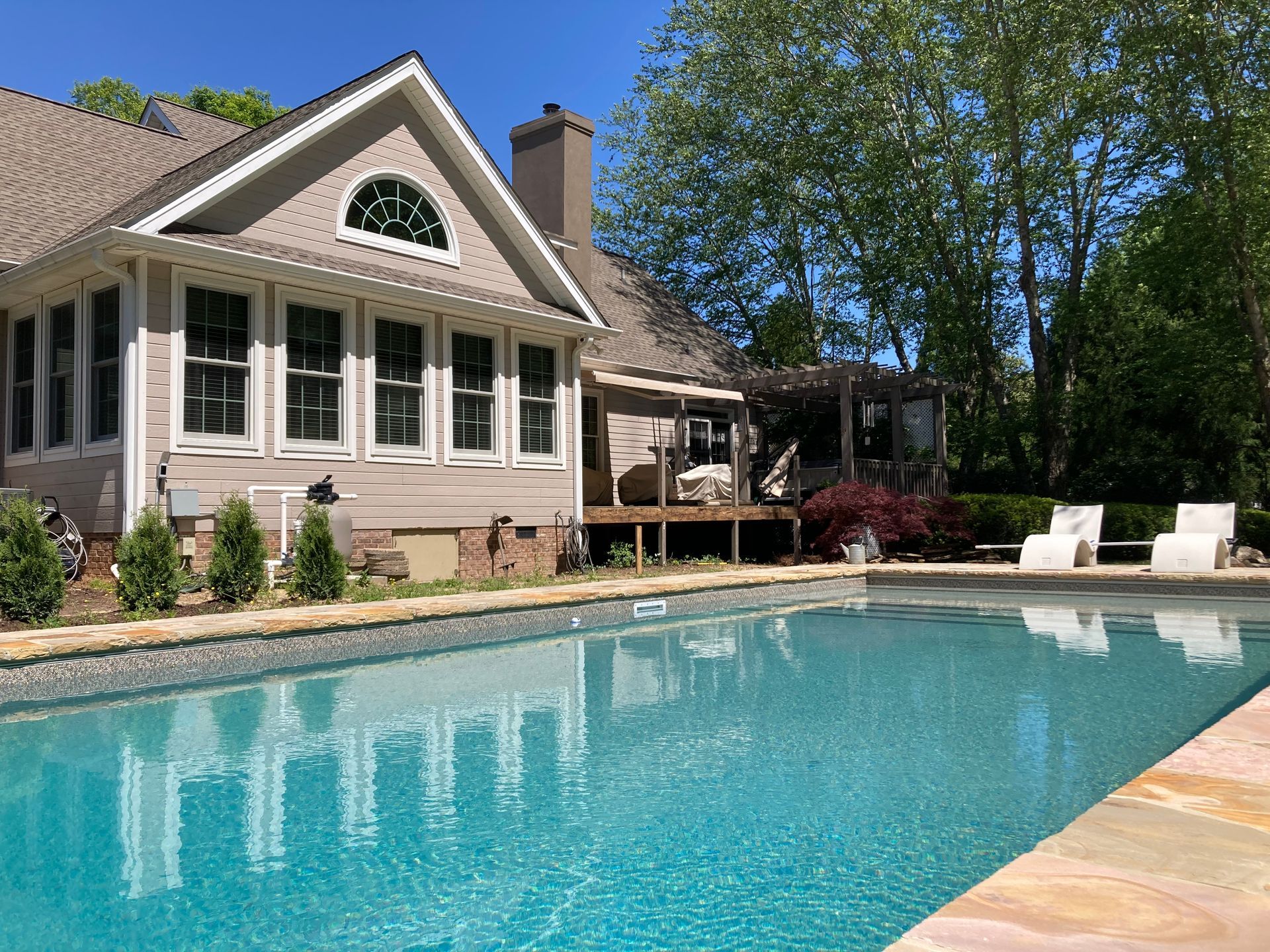 Swimming pool in front of a tan house with a deck. Two white chairs sit near the pool.