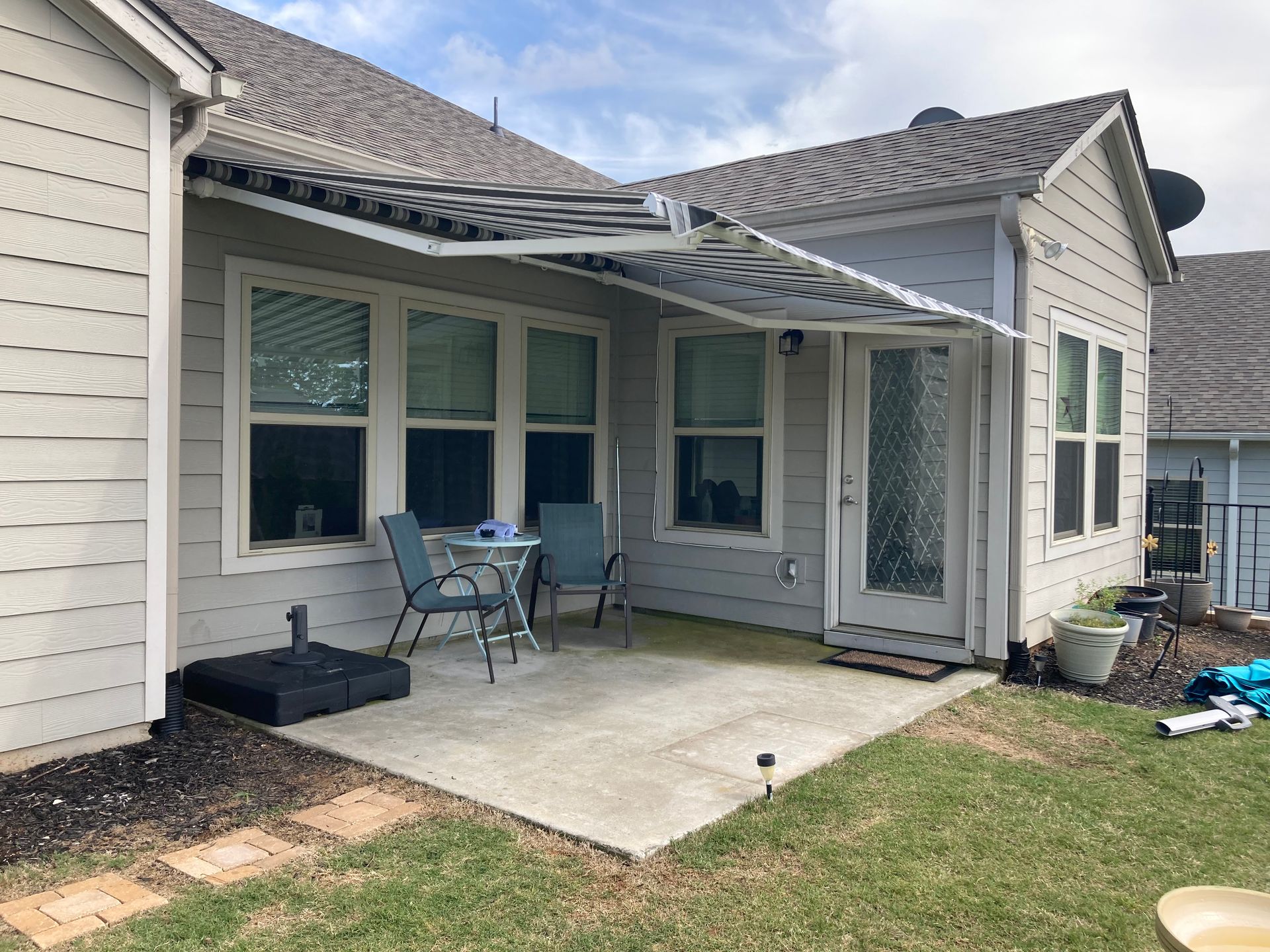 A patio with a retractable awning, chairs, and a small table next to a house with windows and a door.