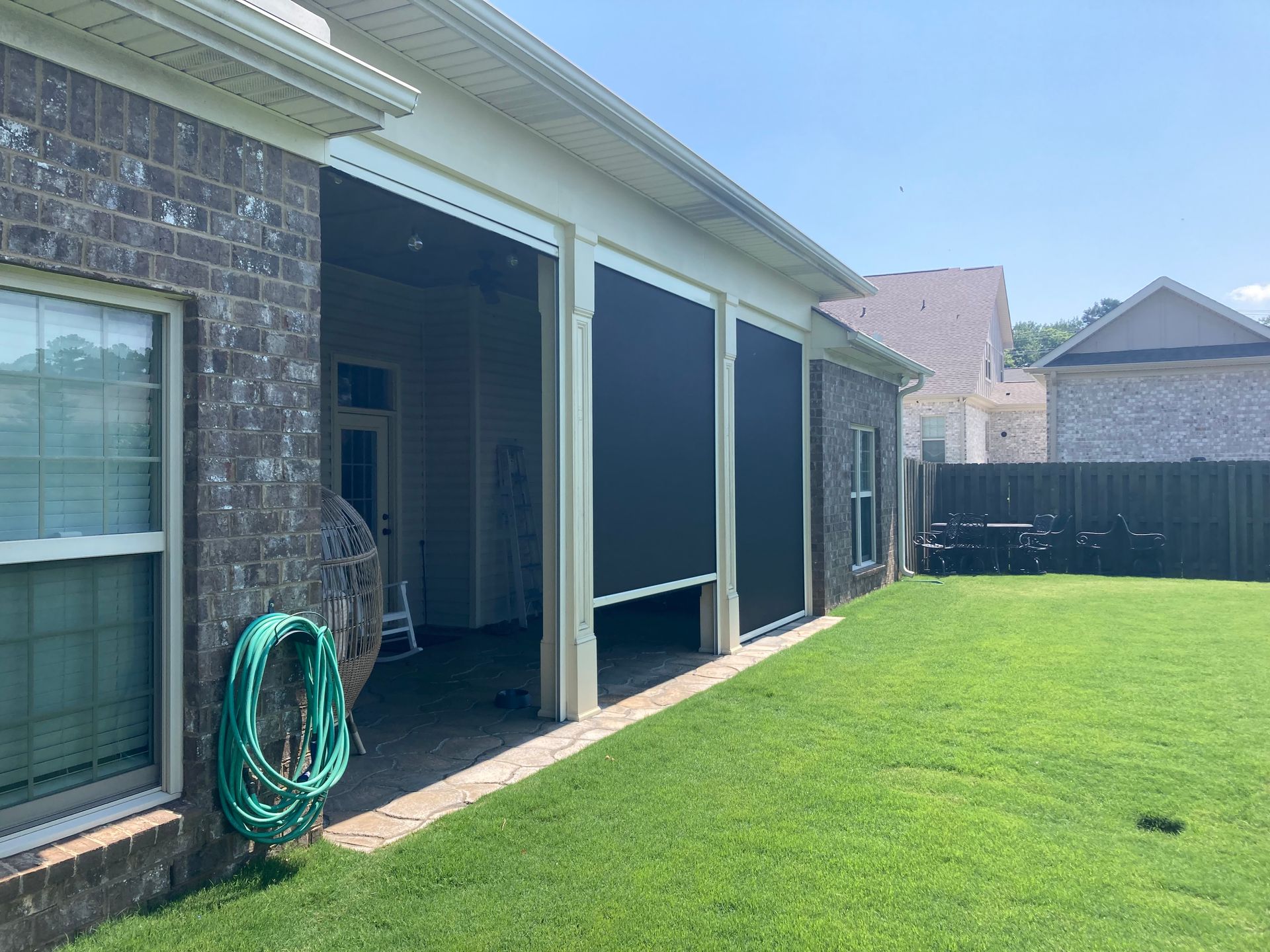 Exterior view of a brick house with retractable black screen shades installed on a patio, overlooking a green lawn.