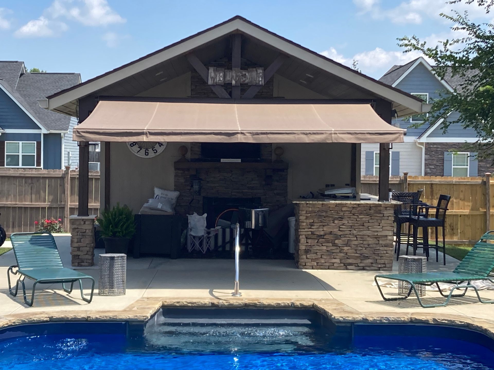 Outdoor poolside structure with a kitchen, fireplace, and bar, shaded by an awning.