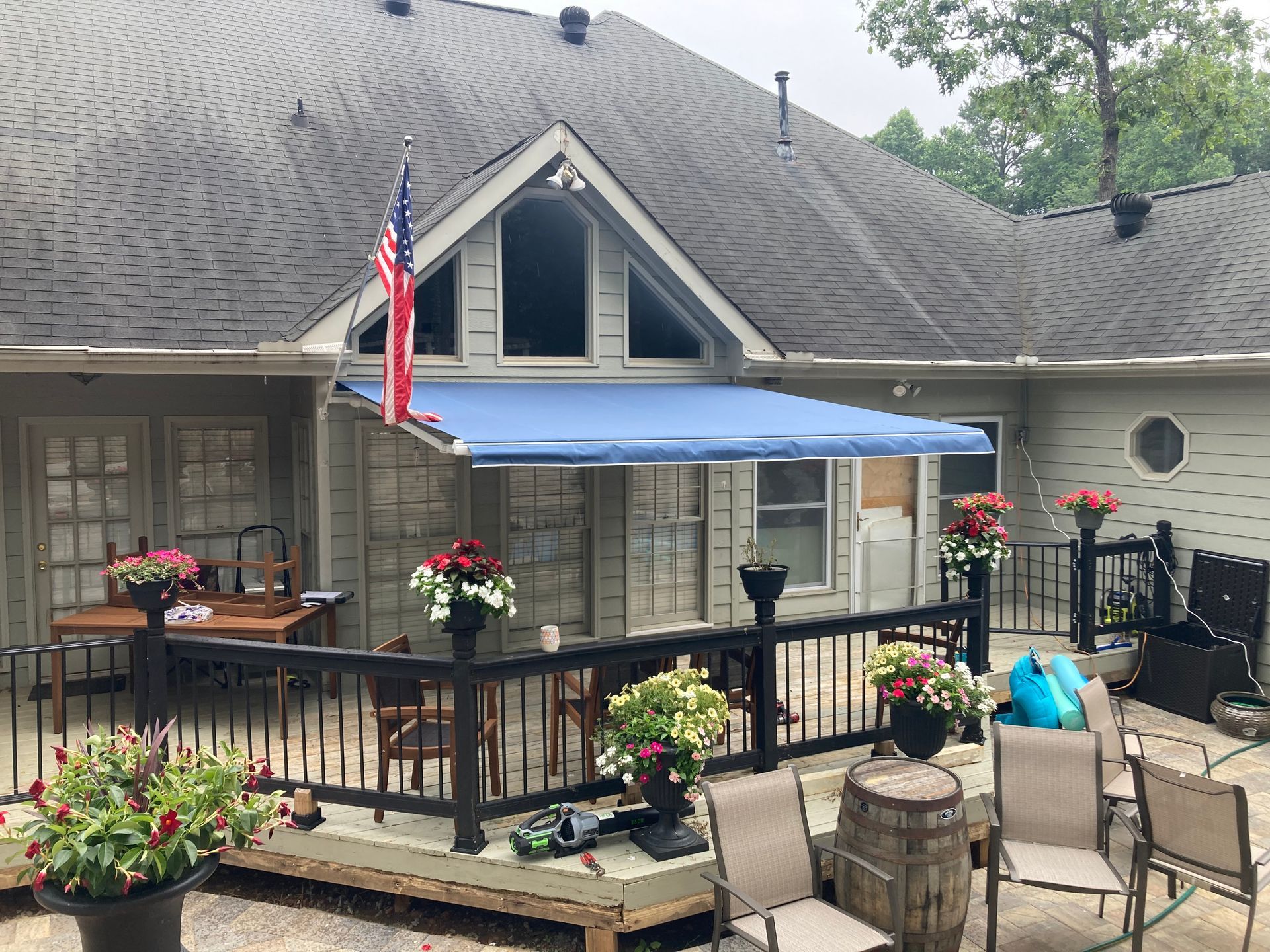 Blue awning over a deck with black railing. American flag and potted flowers visible.