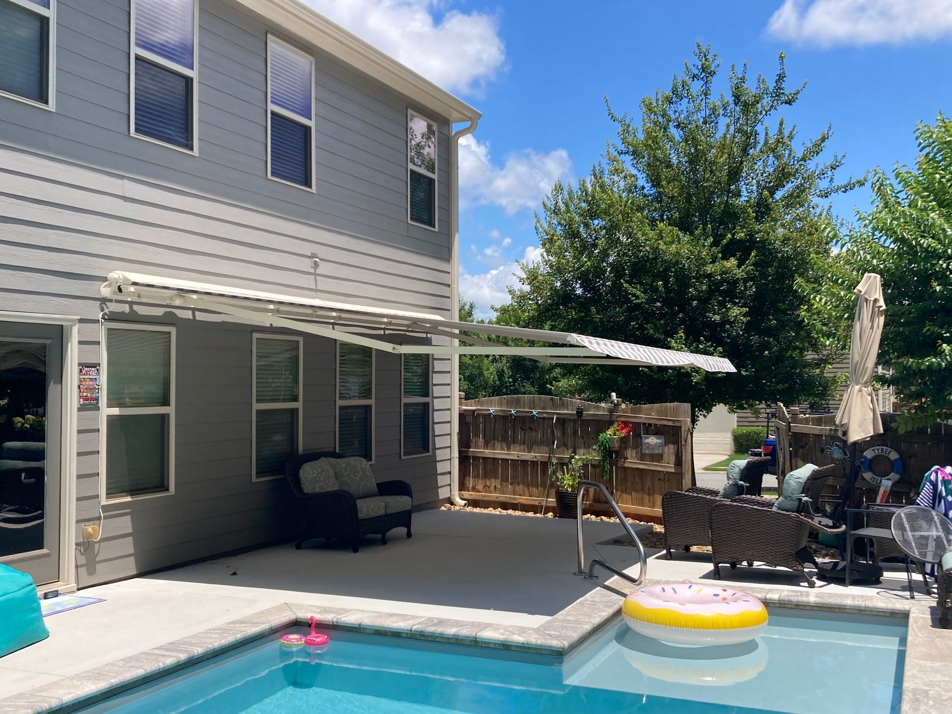 Backyard with a pool, patio, and two-story house. There is an awning, furniture, and a blue sky.