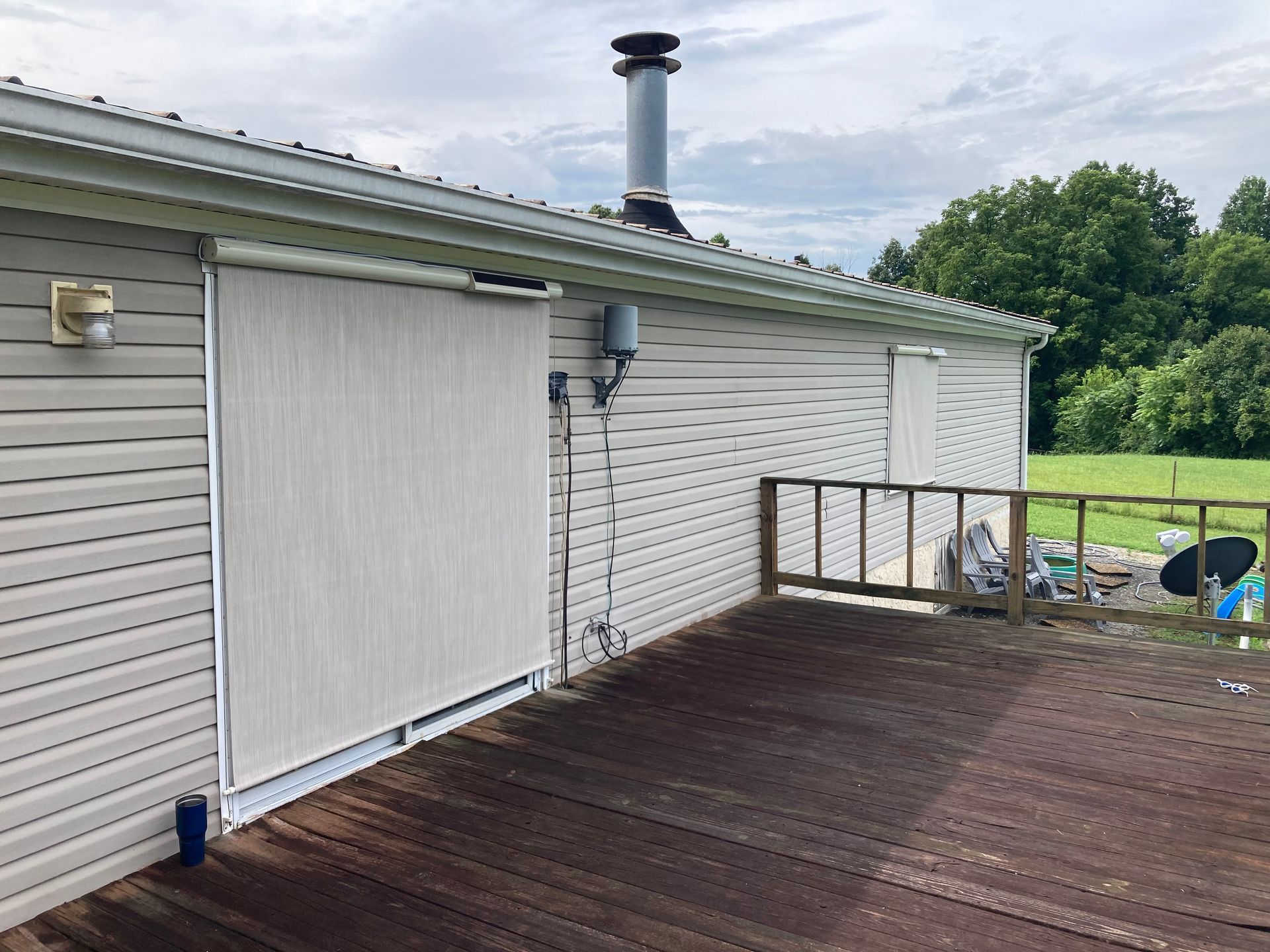 Side of a house with deck and retractable screen shades. Wooden deck, beige siding, trees in background.