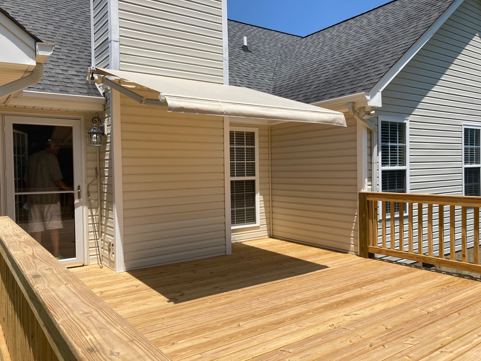 Backyard deck with an awning over a window and door, beige siding, person visible inside door.
