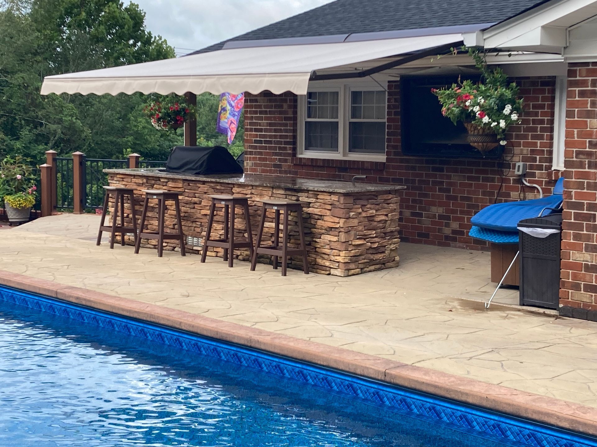 Poolside outdoor kitchen with bar, stools, and retractable awning. Brick and stone facade.
