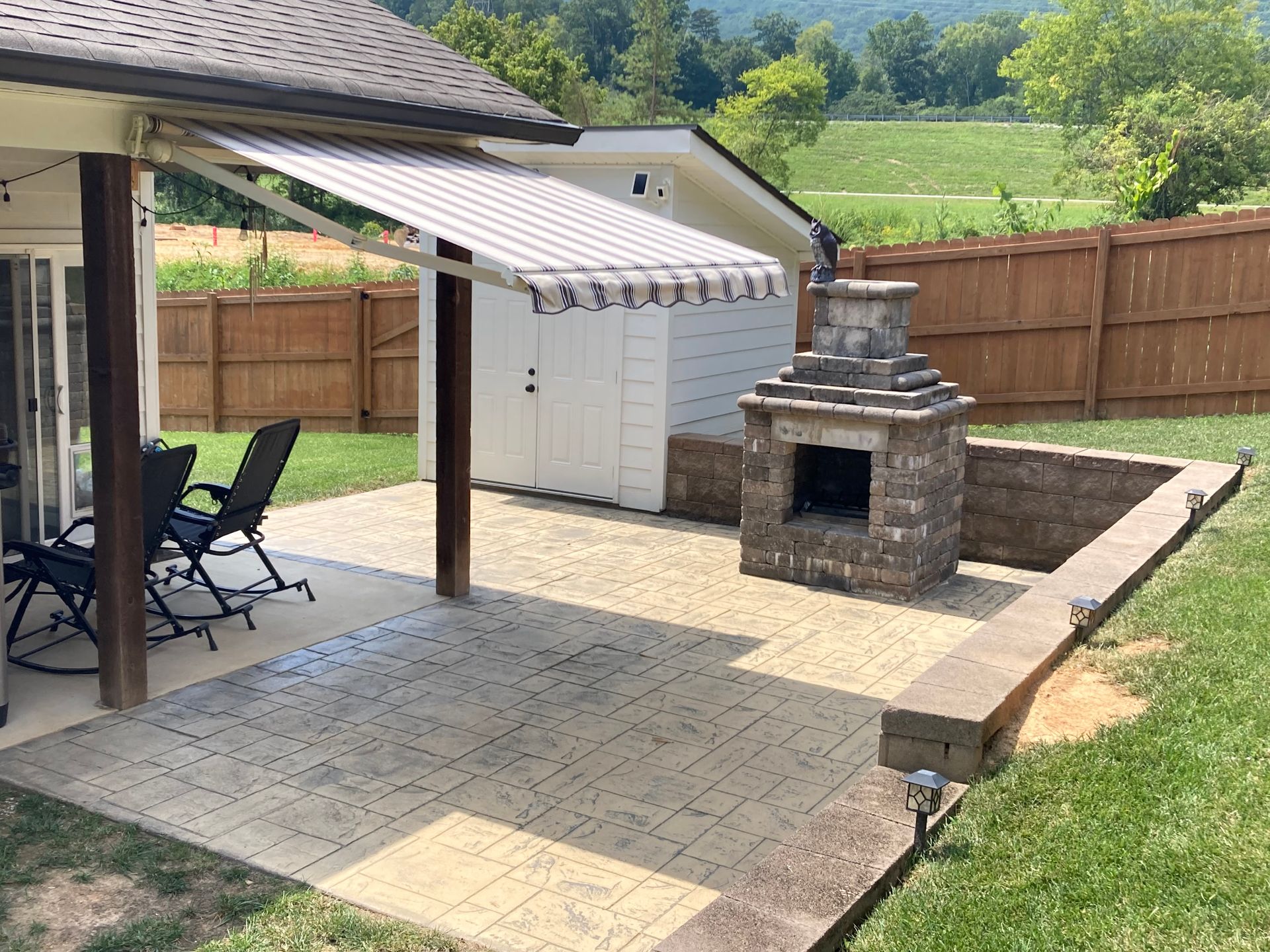 Backyard patio with awning, chairs, fireplace, and shed; surrounded by a fence and grassy area.