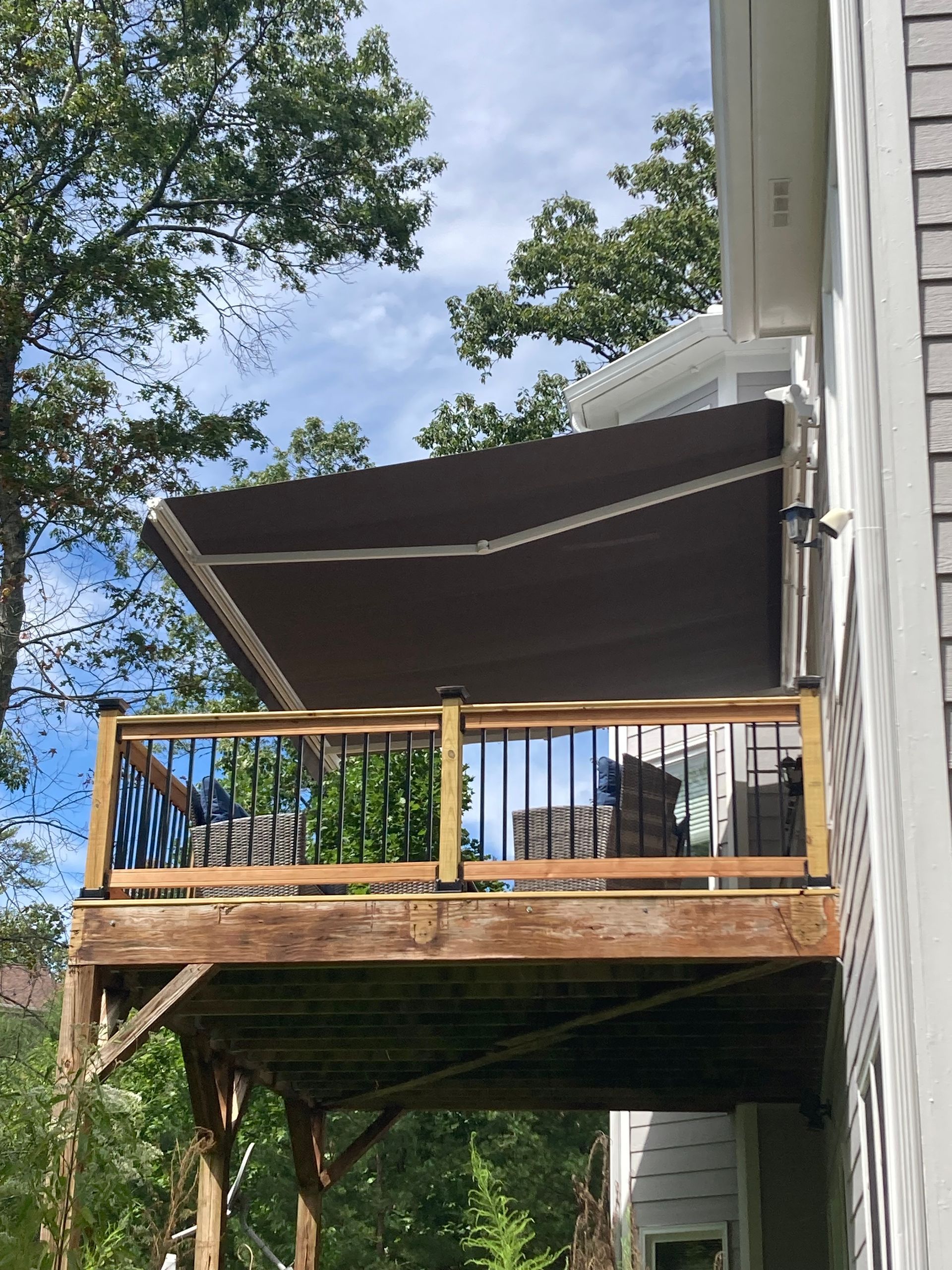 Brown retractable awning over wooden deck with black railings; house exterior on the right; trees in the background.