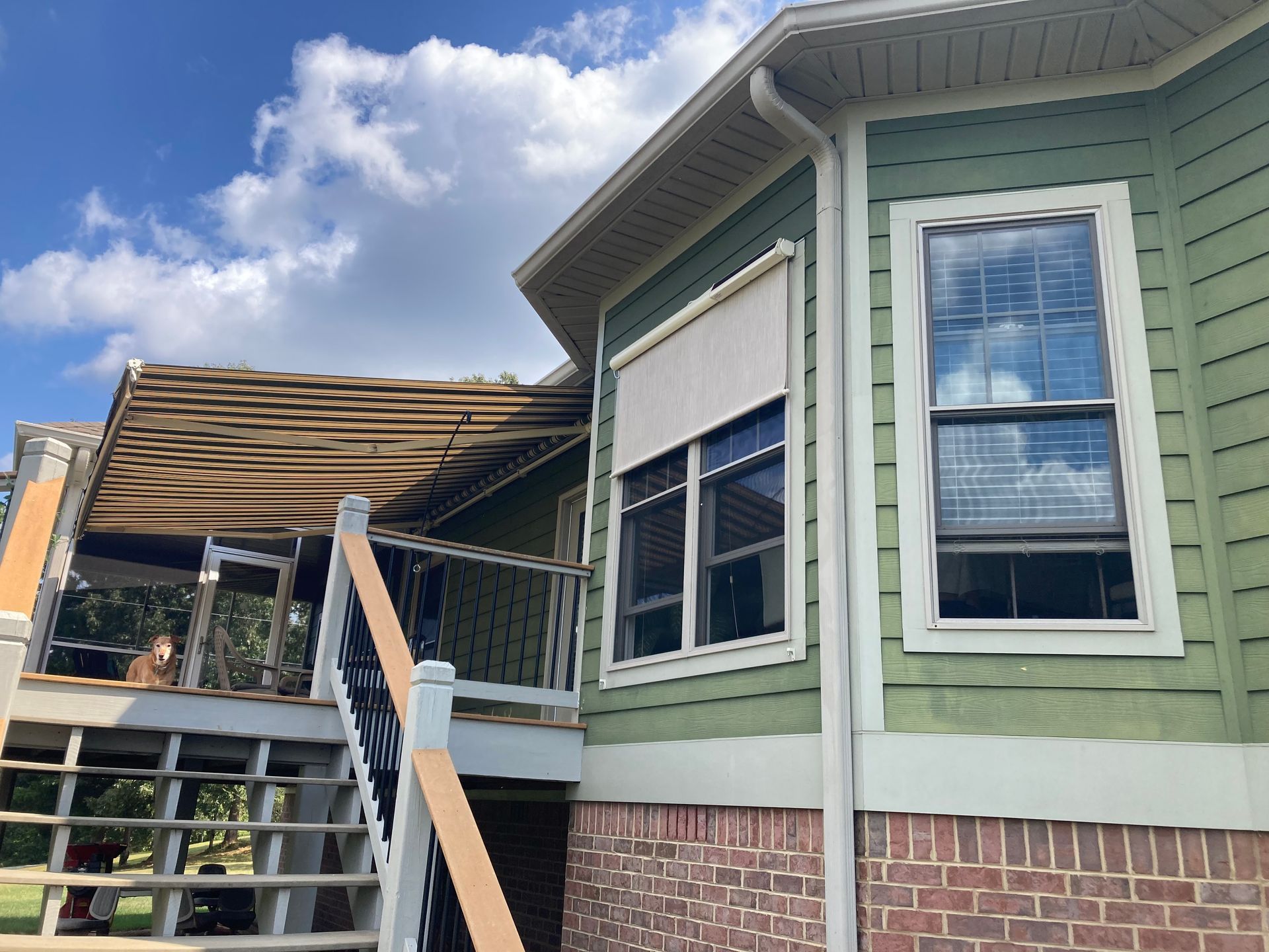 Green house exterior with windows, deck, and a retractable shade. Blue sky with clouds.