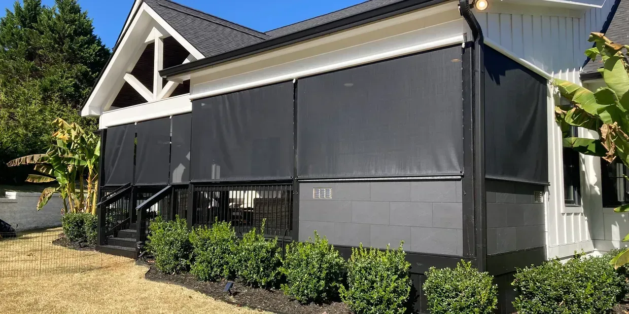 Exterior of a house with black retractable shades on a covered porch. Green shrubbery and grass are visible.