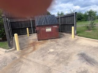 A rusty red dumpster is enclosed by a wooden fence with yellow bollards on a concrete pad. Sky in the background.