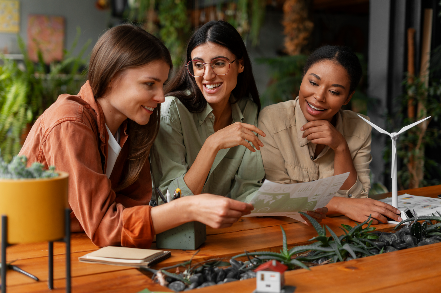 Tres mujeres examinando un plano con una maqueta de casa y un aerogenerador sobre una mesa de madera rodeada de plantas.