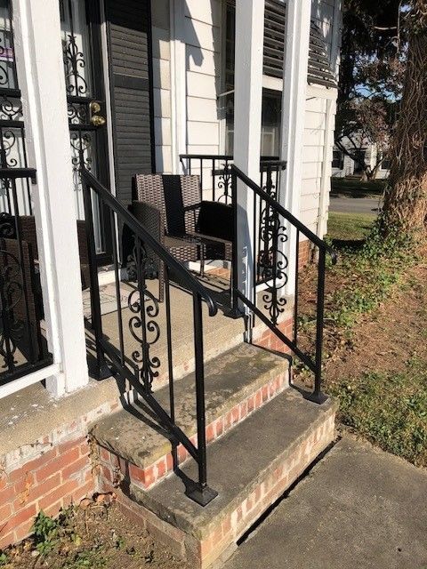 Front porch with a small black metal railing, steps, and a chair beside a house entrance.