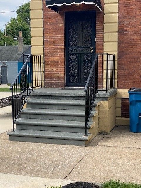 Brick building entrance with steps, black iron railing, and blue trash bin on the right