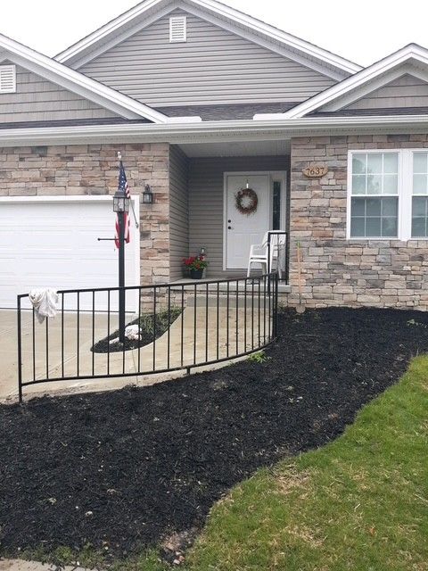 Front porch of a stone house with a ramp, black railing, and mulched flower bed.