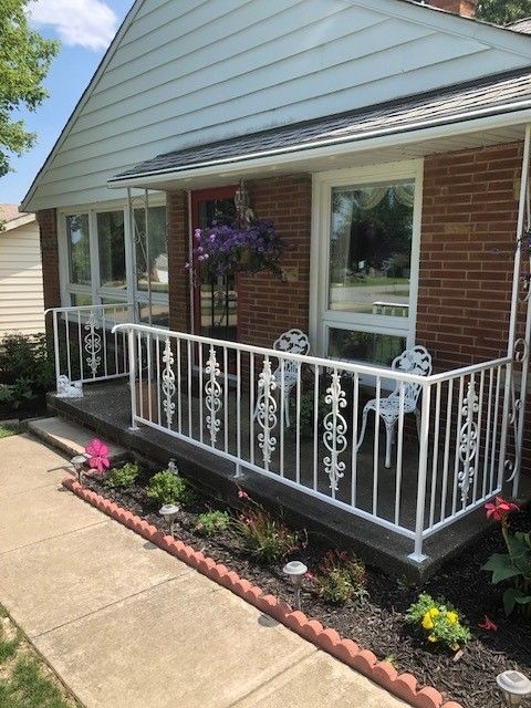 Front porch with white railing, brick house exterior, hanging flowers, and small garden along the walkway