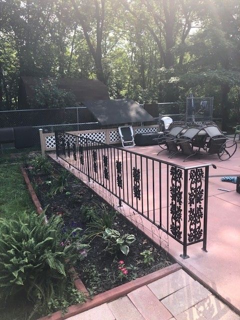 Patio with black wrought-iron railing, lounge chairs, and a grill beside a wooded garden.