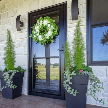 Black front door with a green wreath, flanked by two potted shrubs against a stone facade.