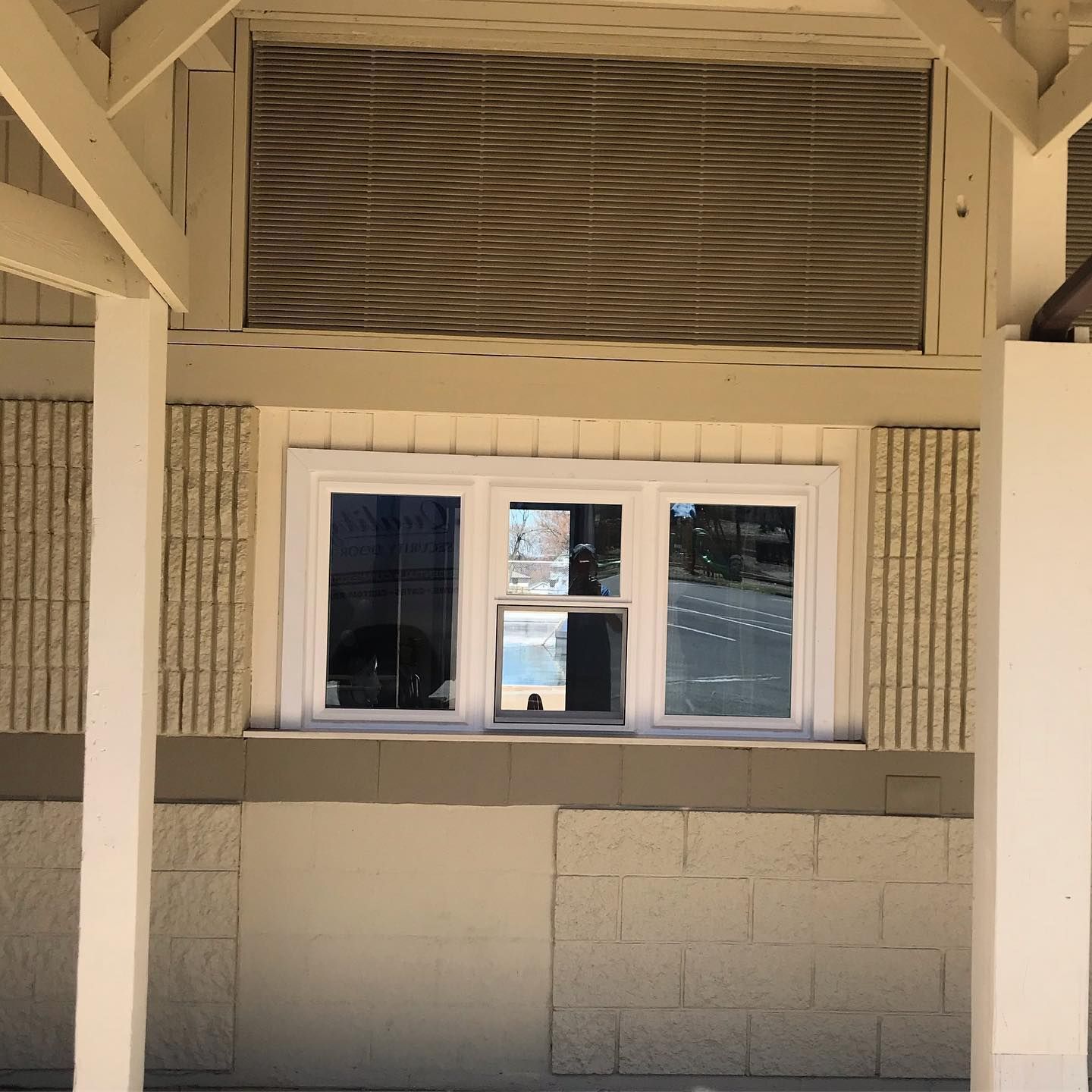 A window with a white frame and a sliding vent is set in a beige wall with textured panels under a covered porch.