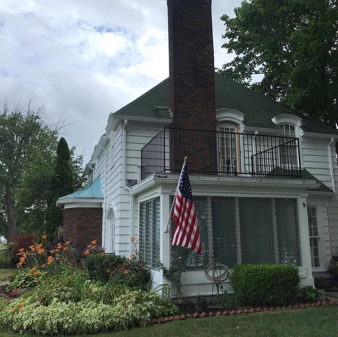 A white, two-story house with a green roof, a large brick chimney, a second-floor balcony, and an American flag.