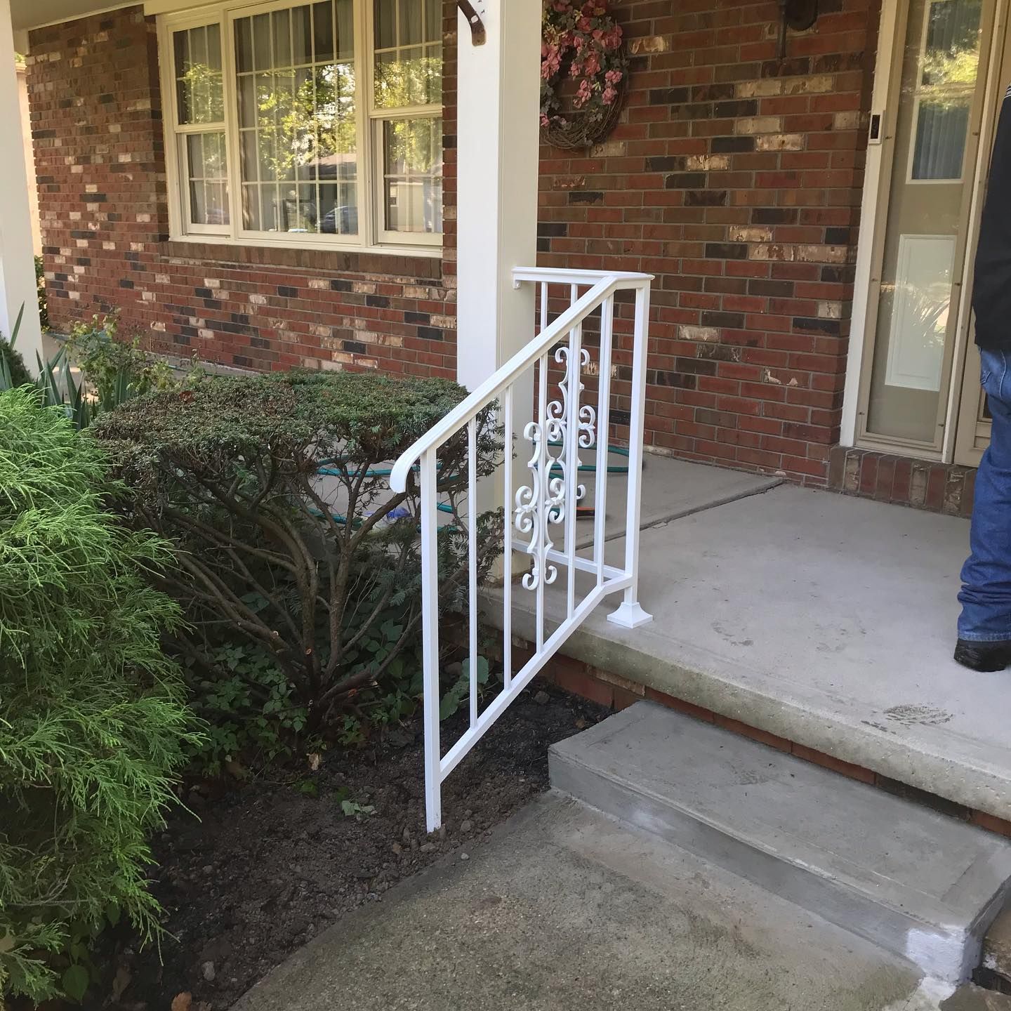 A white metal railing leads up concrete steps to the entrance of a brick house with a front lawn and evergreen bush.