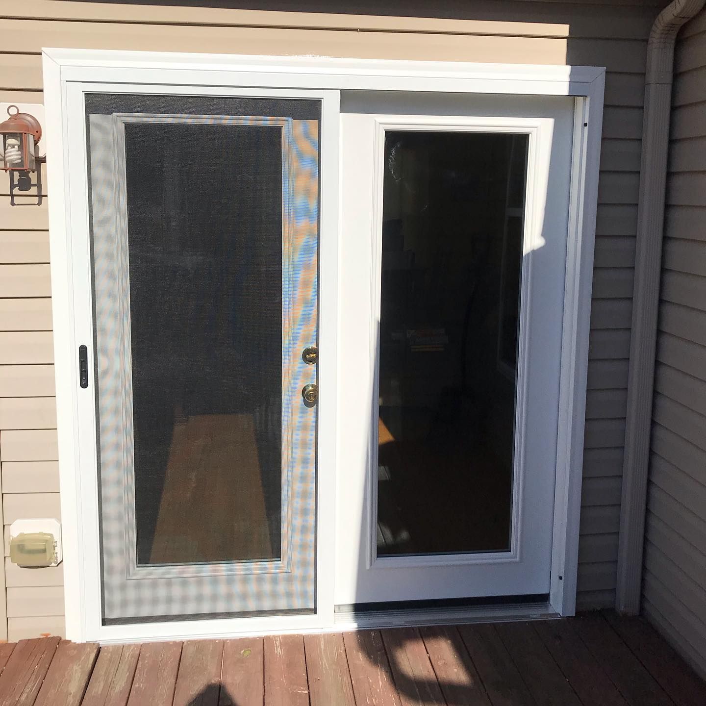 A white-framed exterior sliding door with a black mesh screen door partially open, set against tan horizontal siding.