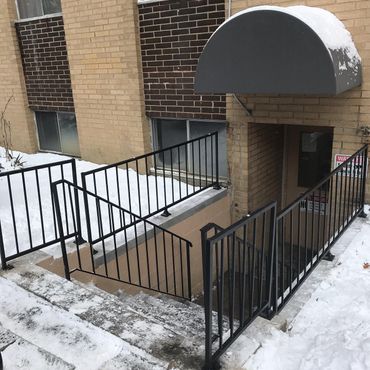 Snow-covered apartment entrance with black metal railings and a gray awning over stairs.
