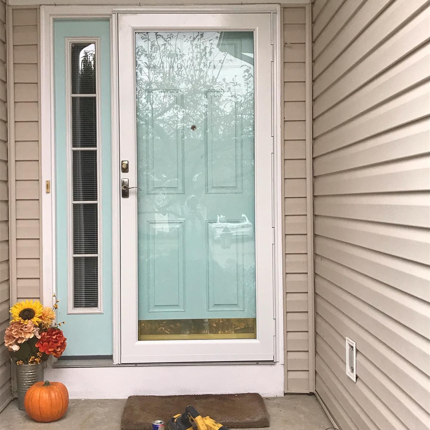 A light blue front door with a windowed side panel, decorated with a small pumpkin and a bouquet of flowers on the porch.