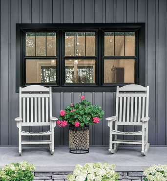 Two white rocking chairs on a porch with a potted pink flower arrangement in front of a black window