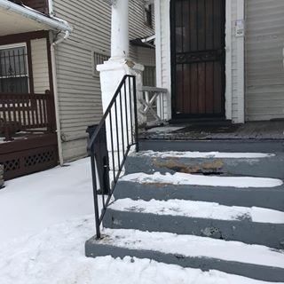 Snow-covered front steps and porch with black railing leading to a house entrance