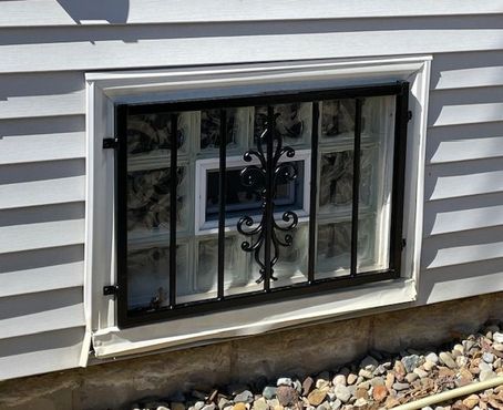 Basement window with black security bars in white vinyl siding above landscaping stones