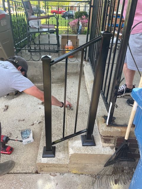 Workers installing a black metal gate section on a concrete walkway beside a porch.