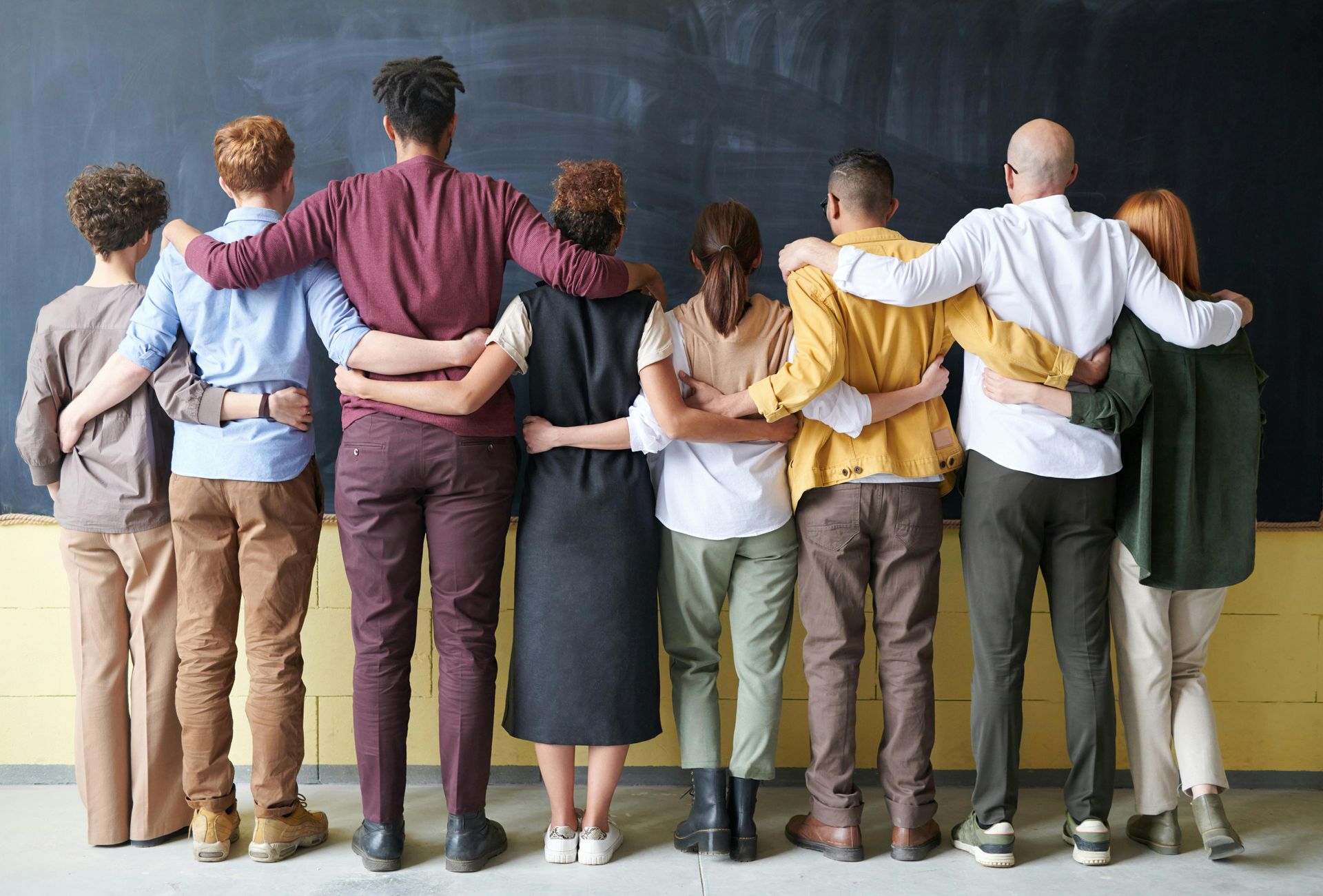 Group of people standing with arms around each other, facing a plain wall in a supportive circle