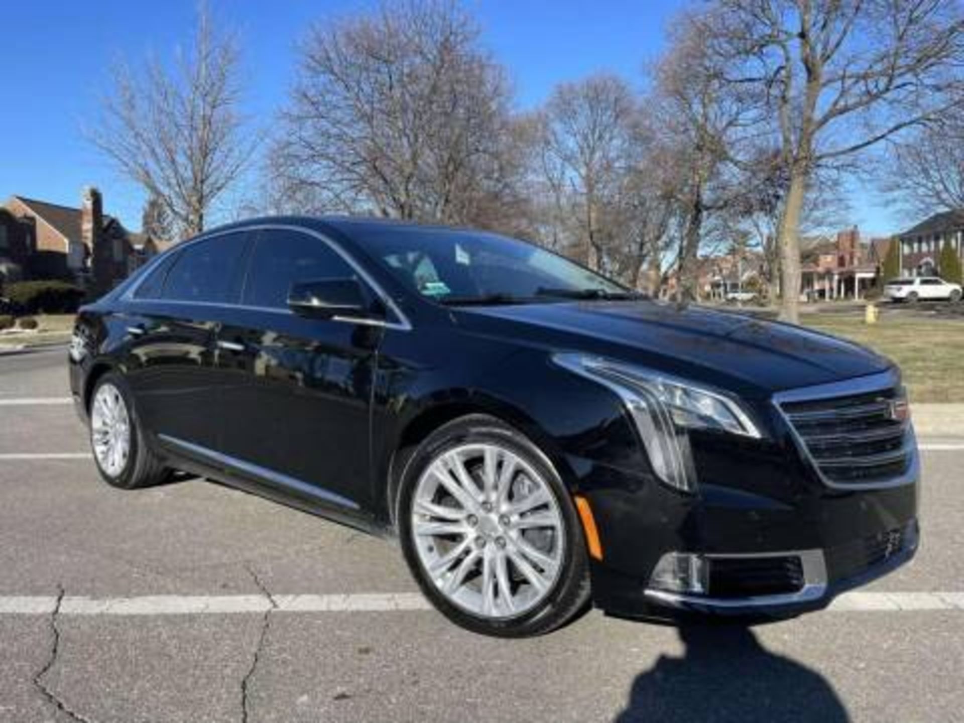 Black Cadillac XTS sedan parked on a street with bare trees and houses in the background on a sunny day.