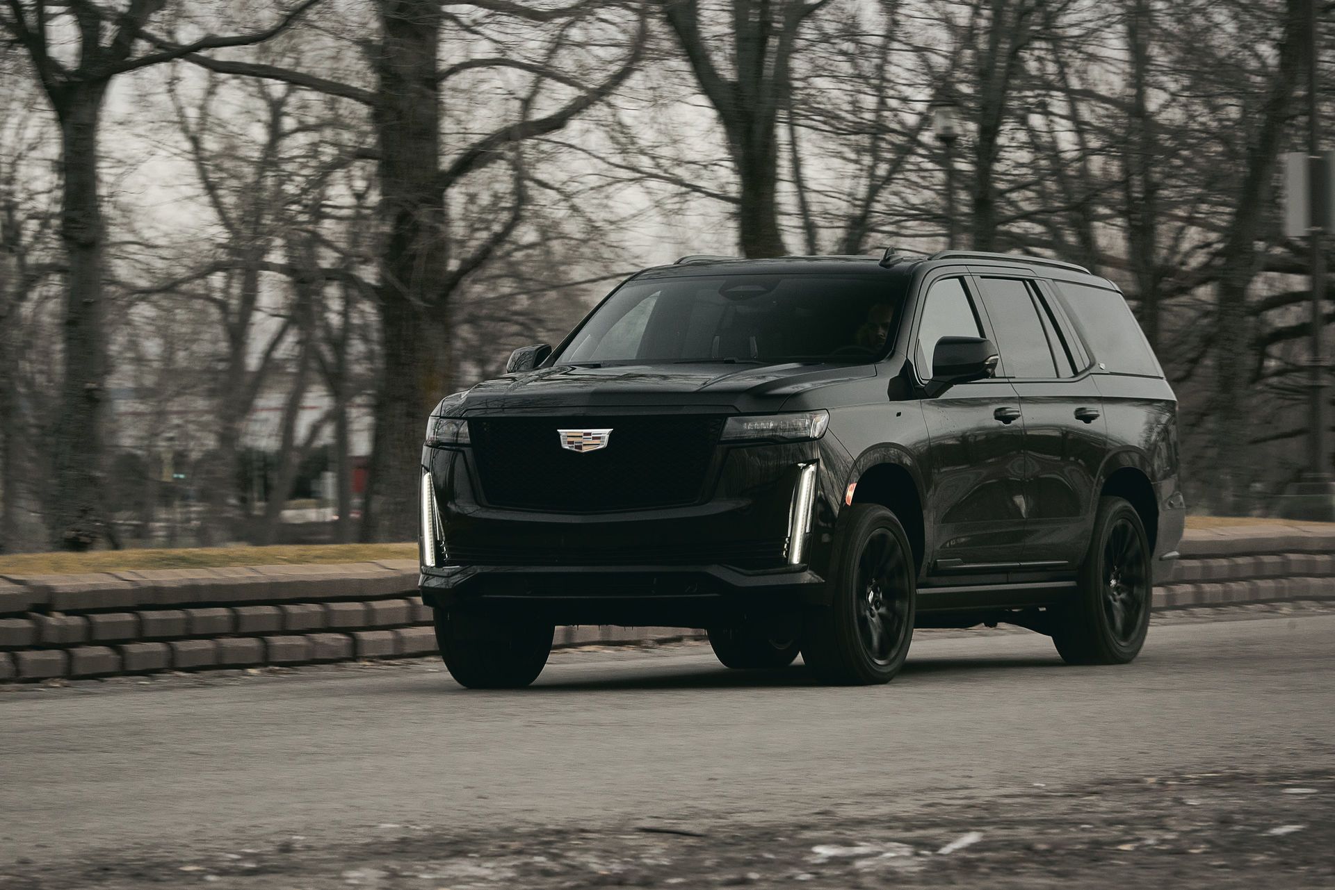 Black Cadillac Escalade SUV driving on a road. It's a cloudy day with trees in the background.